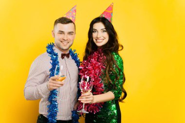 A young couple, a guy and a girl 20-25s in party hats and New Year's tinsel hold glasses of wine or champagne in their hands, smile and look at the camera on a yellow isolated background.