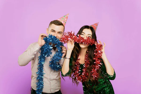 A guy in a bow tie with a girl 20-25s in party hats pose with New Year's tinsel in their hands and look at the camera on a lilac isolated background. Birthday or new year concept