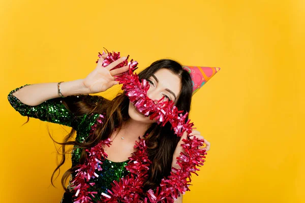 Young beautiful girl 20-25s in a party hat and New Year's tinsel in her hands poses and looks at the camera on a yellow isolated background. Makes an impromptu mustache from New Year's tinsel.