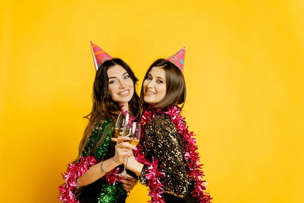 Young adult cheerful girls 20-25s in party hats and New Year's tinsel, hugging holding glasses of wine or champagne and looking at the camera on a yellow isolated background.