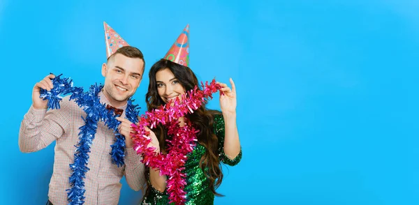 A guy in a bow tie with a girl 20-25s in party hats pose with New Year's tinsel in their hands and look at the camera on a blue isolated background. Party banner with copy space