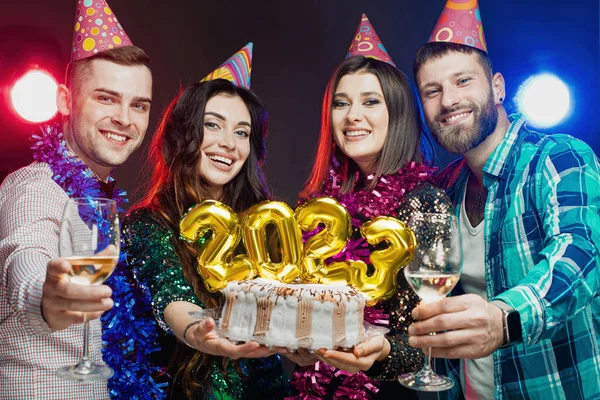 Young adult students in tinsel and party hats hold 2023 number cake at new year party