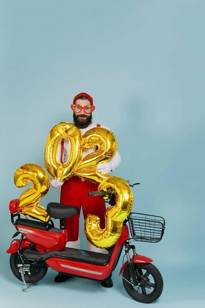 Full length portrait of a hipster man in a santa costume and big heart shaped glasses holding inflatable numbers with the number 2023 in his hands on a red electric scooter on a blue background