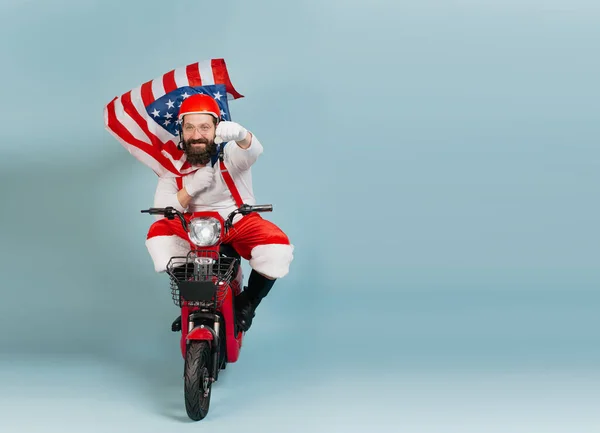 Santa in a superman pose with a fluttering usa flag in the form of a cape and a helmet rides a red electric scooter on a blue isolated background with copy space