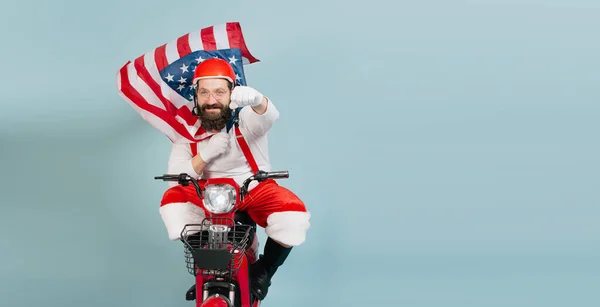 Young adult man in a santa costume in a superman pose with a fluttering usa flag in the form of a cape and a helmet rides a red electric scooter on a blue isolated background with copy space