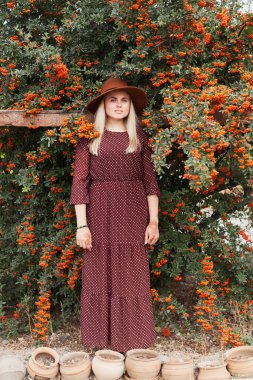 Terracotta kahverengi renkli şapka takan güzel bir kadın ve rowan ağacının yanında olgun turuncu böğürtlenlerle kırsal bir elbise giyiyor. Yavaş yaşam ve doğal güzellik kavramı.