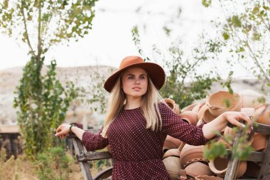 Ahşap kırsal araba dolusu çömlek sürahisinin yanında şapka ve elbise giyen genç bir kadın. Yavaş yaşam tarzı konsepti. Kapadokya Seyahat Maceraları.