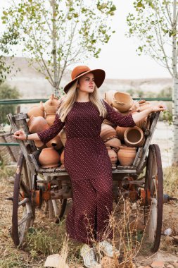 Ahşap kırsal araba dolusu çömlek sürahisinin yanında şapka ve elbise giyen genç bir kadın. Yavaş yaşam tarzı konsepti. Kapadokya Seyahat Maceraları.