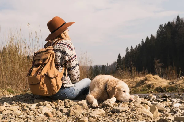 犬と森の小川のそばに腰を下ろして帽子を被った女性バックパッカーが — ストック写真