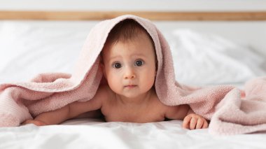 Banyodan ya da duştan sonra ebeveyn yatağında pembe kukuletalı havlu giyen mutlu bir bebek. Bebekler için tekstil. Modern yatak odasında temiz ve kuru bir çocuk. Küçük çocukların yıkanması ve yıkanması. Çocuk hijyeni..