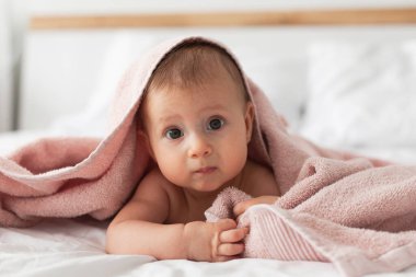 Banyodan ya da duştan sonra ebeveyn yatağında pembe kukuletalı havlu giyen mutlu bir bebek. Bebekler için tekstil. Modern yatak odasında temiz ve kuru bir çocuk. Küçük çocukların yıkanması ve yıkanması. Çocuk hijyeni..