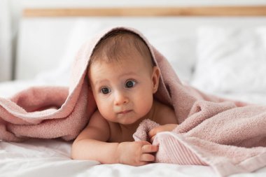 Banyodan ya da duştan sonra ebeveyn yatağında pembe kukuletalı havlu giyen mutlu bir bebek. Bebekler için tekstil. Modern yatak odasında temiz ve kuru bir çocuk. Küçük çocukların yıkanması ve yıkanması. Çocuk hijyeni.. 
