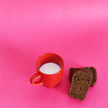 Kırmızı fincanda süt ve pembe arka planda siyah ekmek.