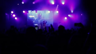 Aydınlatılmış sahne ışıkları. Kaldırılan eller görülebilir. Konser kalabalığı siluetleri