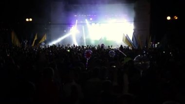 Aydınlatılmış sahne ışıkları. Kaldırılan eller görülebilir. Konser kalabalığı siluetleri