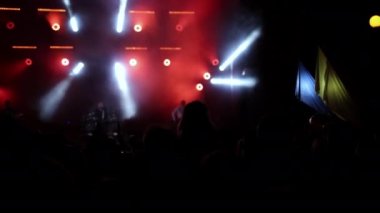 Aydınlatılmış sahne ışıkları. Kaldırılan eller görülebilir. Konser kalabalığı siluetleri