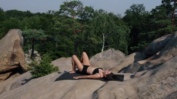 可愛い女はマットの上に寝そべって骨盤 足を上げてる 美しい女性は岩の上で朝の間にワークアウトをする — ストック動画
