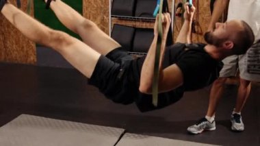 Antrenör monitörleri egzersizi. Çok çalışıyorum. Genç atletik bir adamın yan görüntüsü spor kıyafetleri içinde spor salonunda trx fitness kayışlarıyla havada asılı duran mükemmel bir vücut. Erkekler spor salonunda kollarını jimnastik kemerleriyle çalıştırıyorlar.