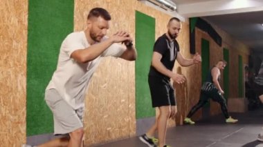 Sportif, mutlu genç erkekler ısınıyor, sabah koşusundan önce esniyorlar, yüksek hamleler yapıyorlar, bacaklar, kalçalar ve kalçalar için egzersiz yapıyorlar, kapalı alanda çalışıyorlar. Tam uzunluk