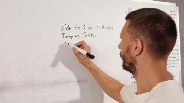 安排和计划你的锻炼 一个穿着T恤的人在白板上写下了一个训练计划 培训课 培训课 — 图库视频影像