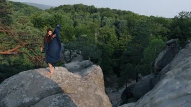 Mavi elbiseli, saçları tüylü vahşi ortaçağ kadını uçurumun kenarında güneşe bakıyor. Dans eder, sallanan elbiseler giyer ormanın arkasında