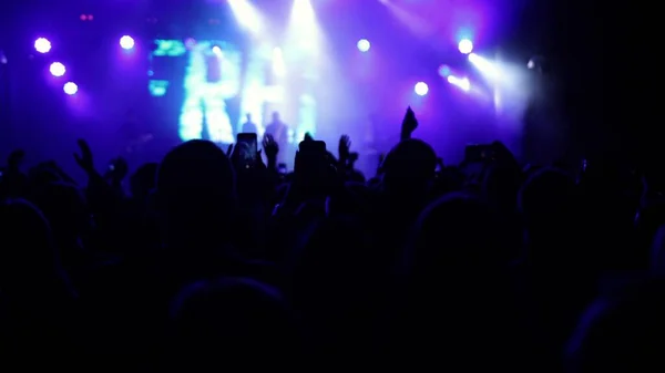 Illuminated by stage lights. Raised hands are visible. Concert crowd silhouettes in front of bright stage. People with raised hands. Concert crowd silhouettes in front of bright platform lights