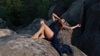 There is a young woman sitting on rocky terrain, relaxing outdoors while wearing a blue silk dress while enjoying a travel lifestyle in the background of mountains