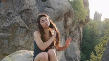 young woman relaxing outside in a blue silk dress sitting on rocky terrain with mountains in the background, relaxing in the outdoors as part of a travel lifestyle