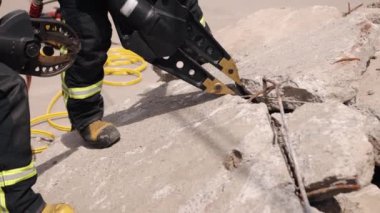 There is a man in a uniform rescuing a victim of an earthquake, using a power hydraulic cutting tool, hydraulic clamps, and is a firefighter, as well