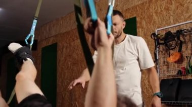 Koç nasıl kaldırılacağını gösteriyor. Çok çalışıyorum. Genç atletik bir adamın yan görüntüsü spor kıyafetleri içinde spor salonunda trx fitness kayışlarıyla havada asılı duran mükemmel bir vücut. Erkekler spor salonunda kollarını trx fitness kayışlarıyla çalıştırıyor.