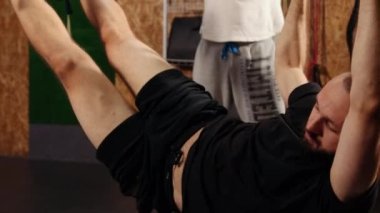 Working hard. Side view of young athletic man, perfect body in sportswear hanging in air with trx fitness straps in gym. Men training arms with trx fitness straps in gym. Trainer monitors exercise.