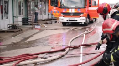 A group of firefighters are preparing to start one of the crews while wearing protective gear with helmets on, a fire engine is visible in the background.