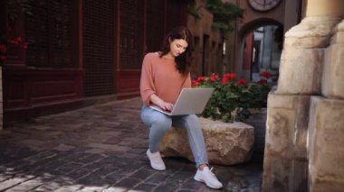 Female typing on laptop keyboard whilst sitting on decorative stone of city streets, attractive young woman using laptop, pretty girl preparing for classes in nature, woman studying outside