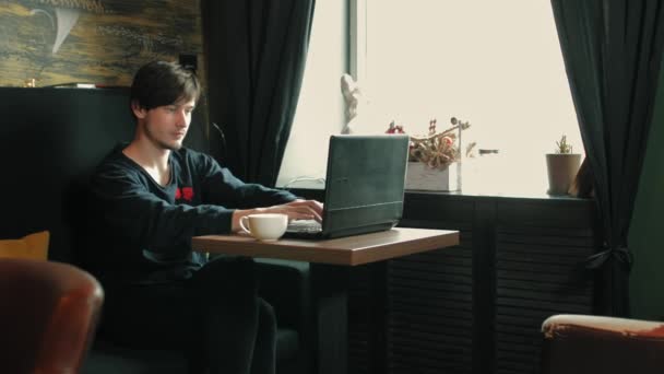 Young freelance guy works at a laptop in a morning cafe and drinks coffee — Video Stock