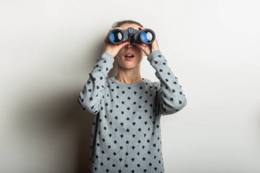 Süveter giymiş, şaşırmış yüzlü genç bir kadın arka plandaki dürbünle bakar..