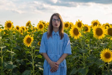 Gün batımında ayçiçeği tarlasında giysili ve gözlüklü genç bir kadın..