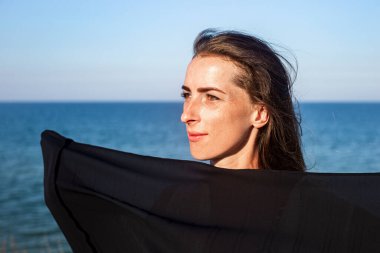 Güzel genç bir kadın, vücudunu denizin ve gökyüzünün arka planına karşı siyah bir bezle örtüyor. 