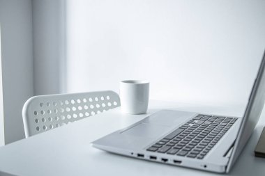 Laptop, beyaz bardak, beyaz bir masada kapalı bir çiçek. İşyeri 