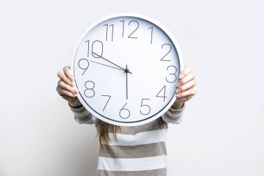 Kadınların elleri beyaz bir duvar saati tutuyor. Yüzler arka planda görünmüyor. 