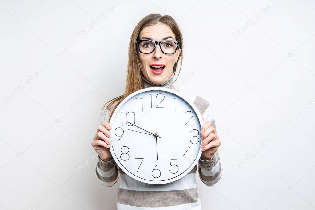 Surprised young woman holding white wall clock on light background 