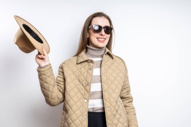 Gözlüklü ve ceketli gülümseyen genç bir kadın beyaz bir duvarın yanında şapkasını çıkardı.