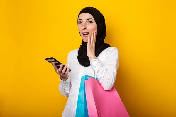 Zaskoczona Młoda Muzułmanka Hidżabie Trzyma Torby Zakupy Telefon Żółtym Tle — Zdjęcie stockowe