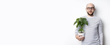 Gözlüklü sakallı bir adam bir saksıda çiçek tutuyor ve beyaz bir duvara yaslanıyor..