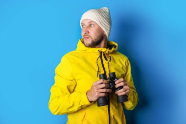 Dürbünlü sarı ceketli genç adam mavi arka planda yan tarafa bakıyor..