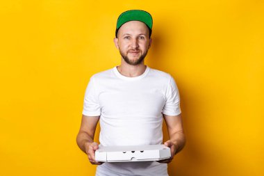 Beyzbol şapkalı gülümseyen genç adam sarı arka planda bir paket pizza tutuyor.. 