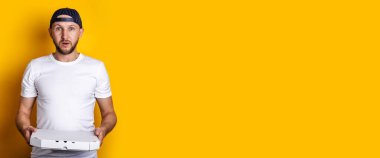 Sarı arka planda paketlenmiş pizza tutan beysbol şapkalı genç adam şaşırdı. Pankart.