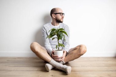 Genç adam, elinde çiçek saksısı tutarken tahta zeminde oturuyor.