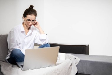 Dizüstü bilgisayarı olan beyaz gömlekli genç bir kadın kanepede otururken çalışıyor.