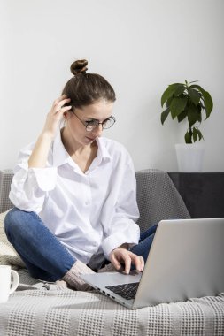 Dizüstü bilgisayarı olan beyaz gömlekli genç bir kadın kanepede otururken çalışıyor.