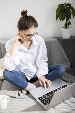 Dizüstü bilgisayarı olan beyaz gömlekli genç bir kadın kanepede otururken çalışıyor.